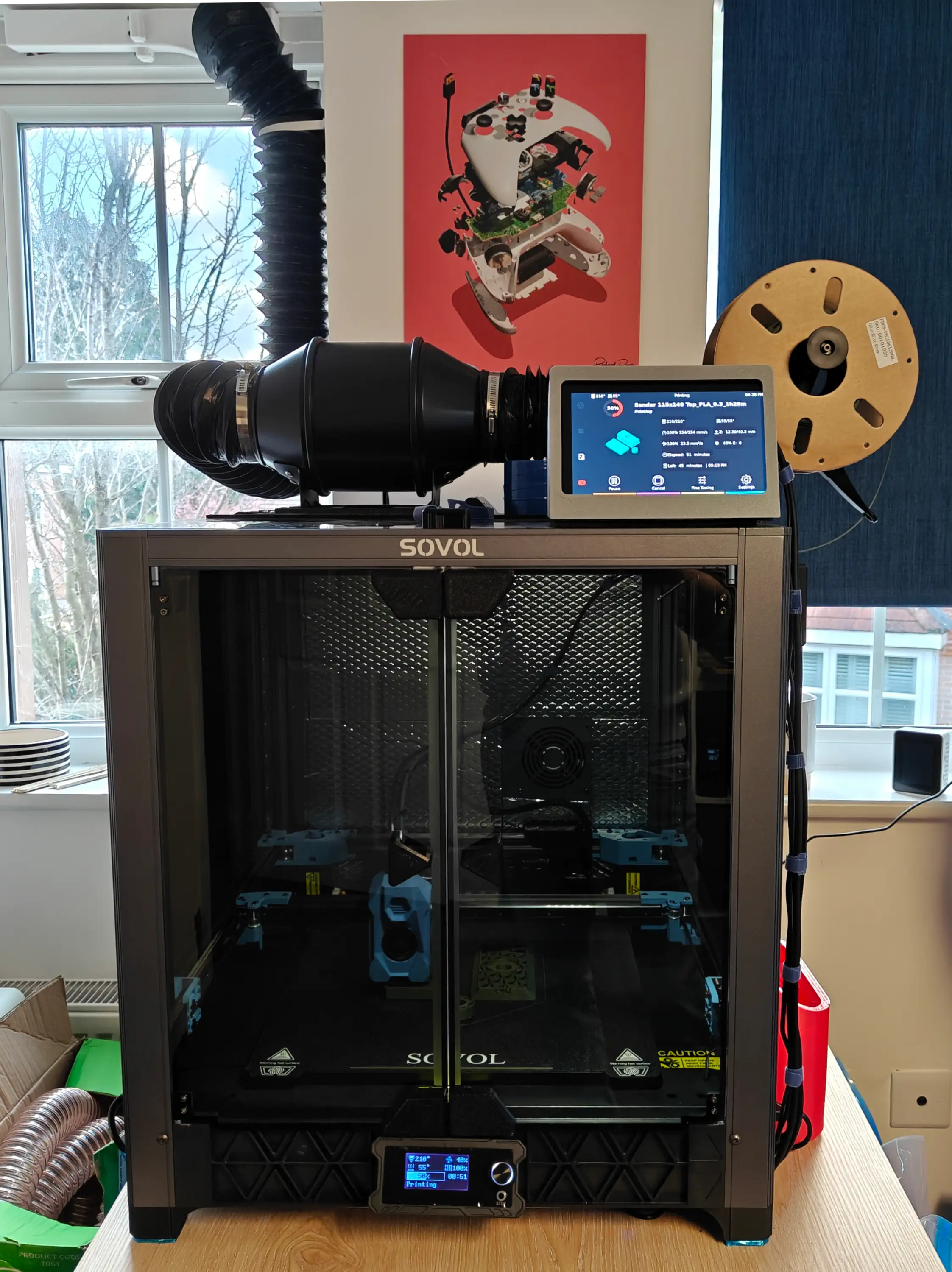 Front on view of the printer, a large silver box with dark tinted front glass doors with the print head active behind it. A screen sits on top along with some tubing running into a fan then up along to the top of a window