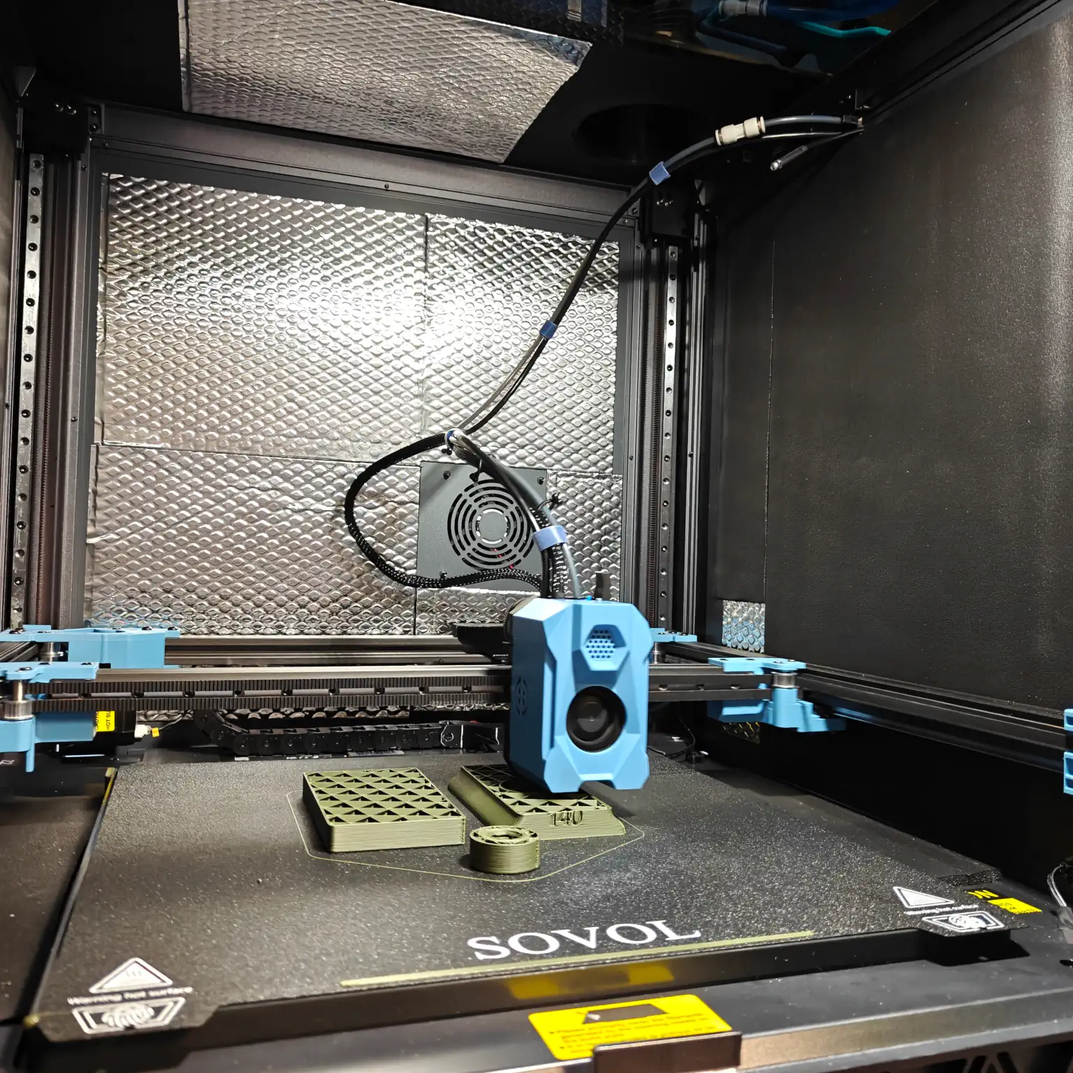A view inside the printer, with a blue print head on a frame gantry, printing some green parts. A couple of the interior panels are shiny silver, with others black.