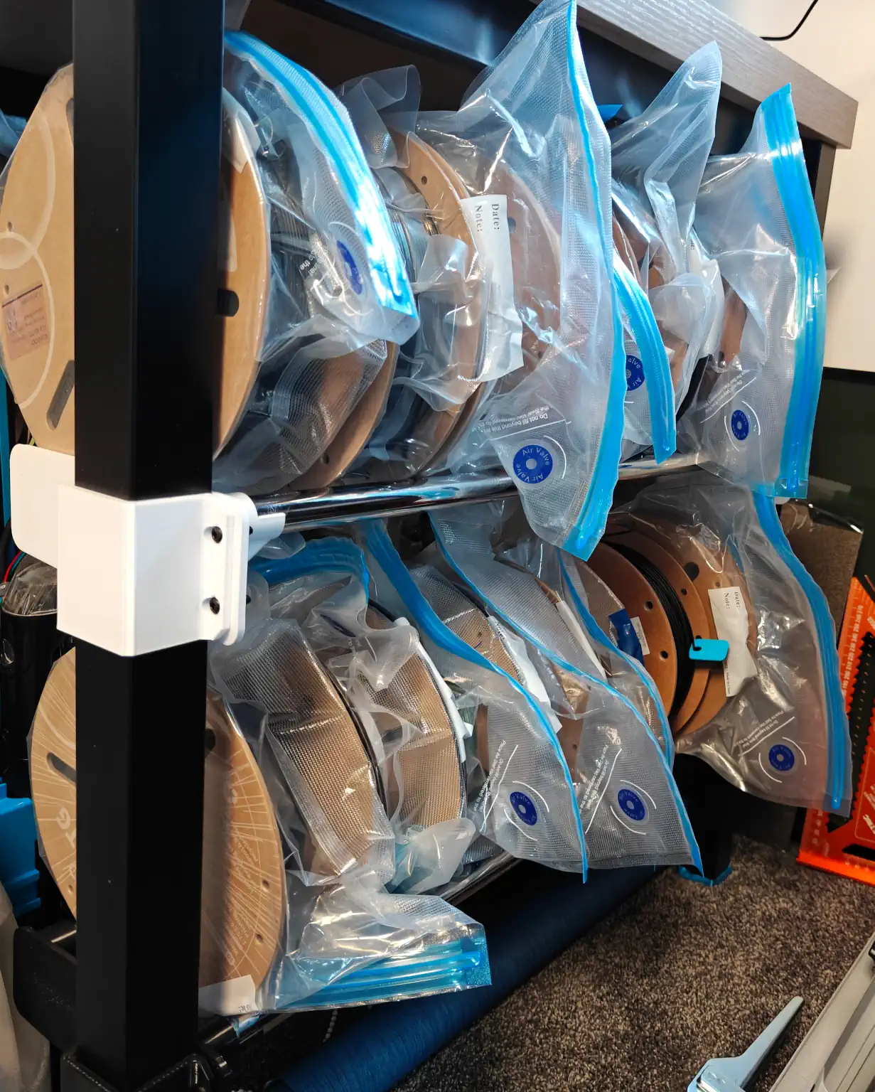 Two rows of shelves, with metal bar bases, running alongside the side of a table, clamped to the legs. Each shelf is full of filament spools