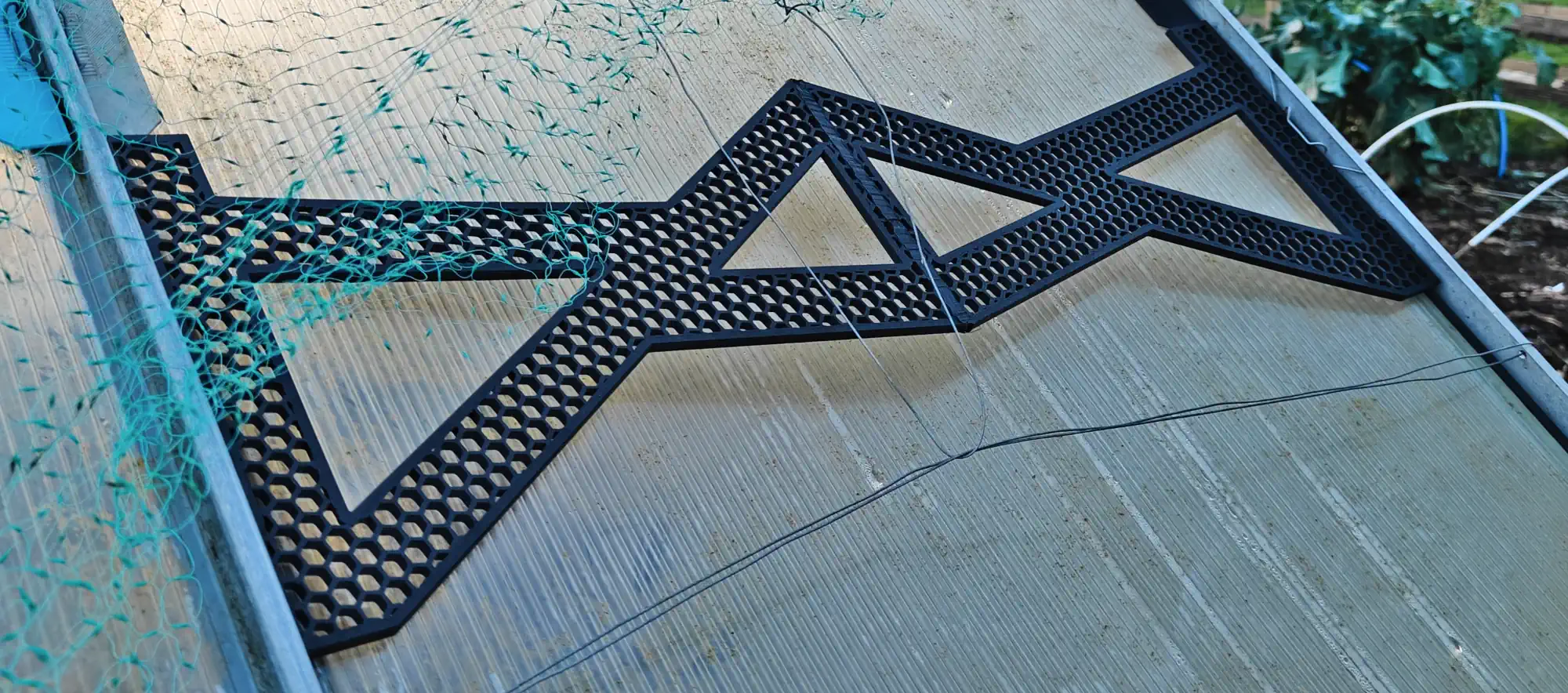 A black plastic part sitting in front of a greenhouse roof panel, fixed on either side. It has a hexagonal pattern of holes in it, within a triangular design.