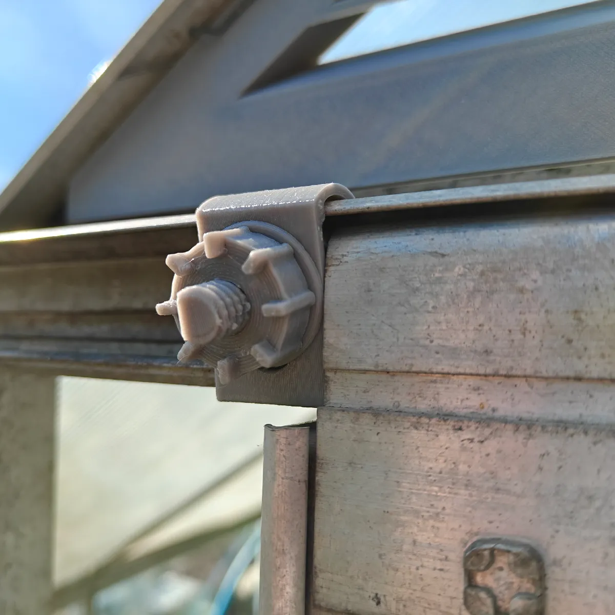 A close view of a greenhouse door rail, showing a grey plastic part butting up against the door, which wraps the rail and appears to have a nut on the front, with a bolt going through it