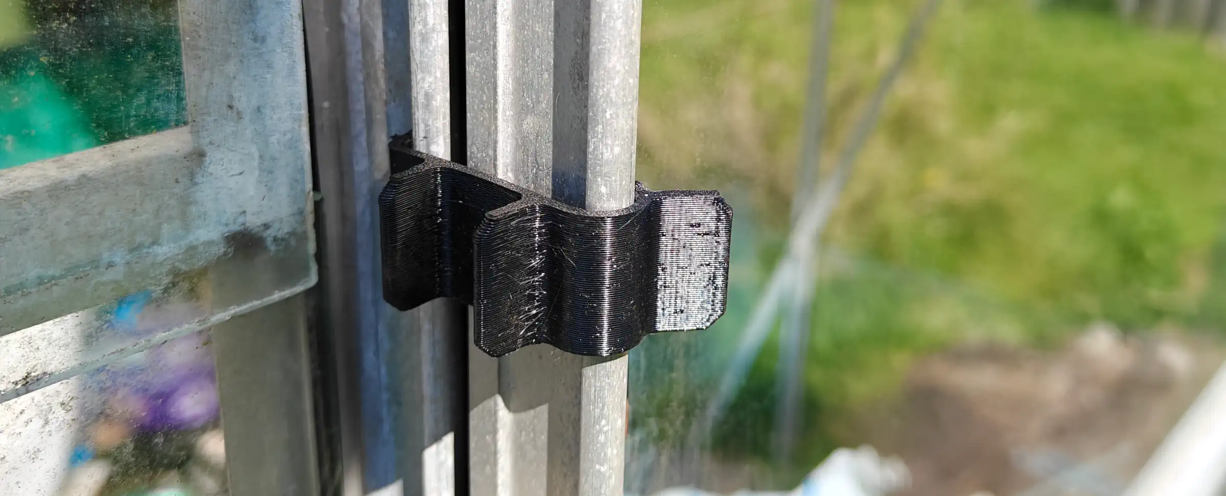 A black plastic part hooked around the edges of a greenhouse door frame, and the edge of the door itself. There&rsquo;s little tabs along the side of it.
