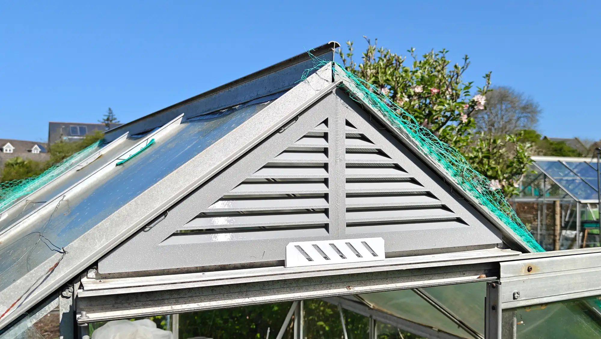 A view of the front of a greenhouse, above the door, with a grey plastic triangular part, with vent slats, sat within the frame