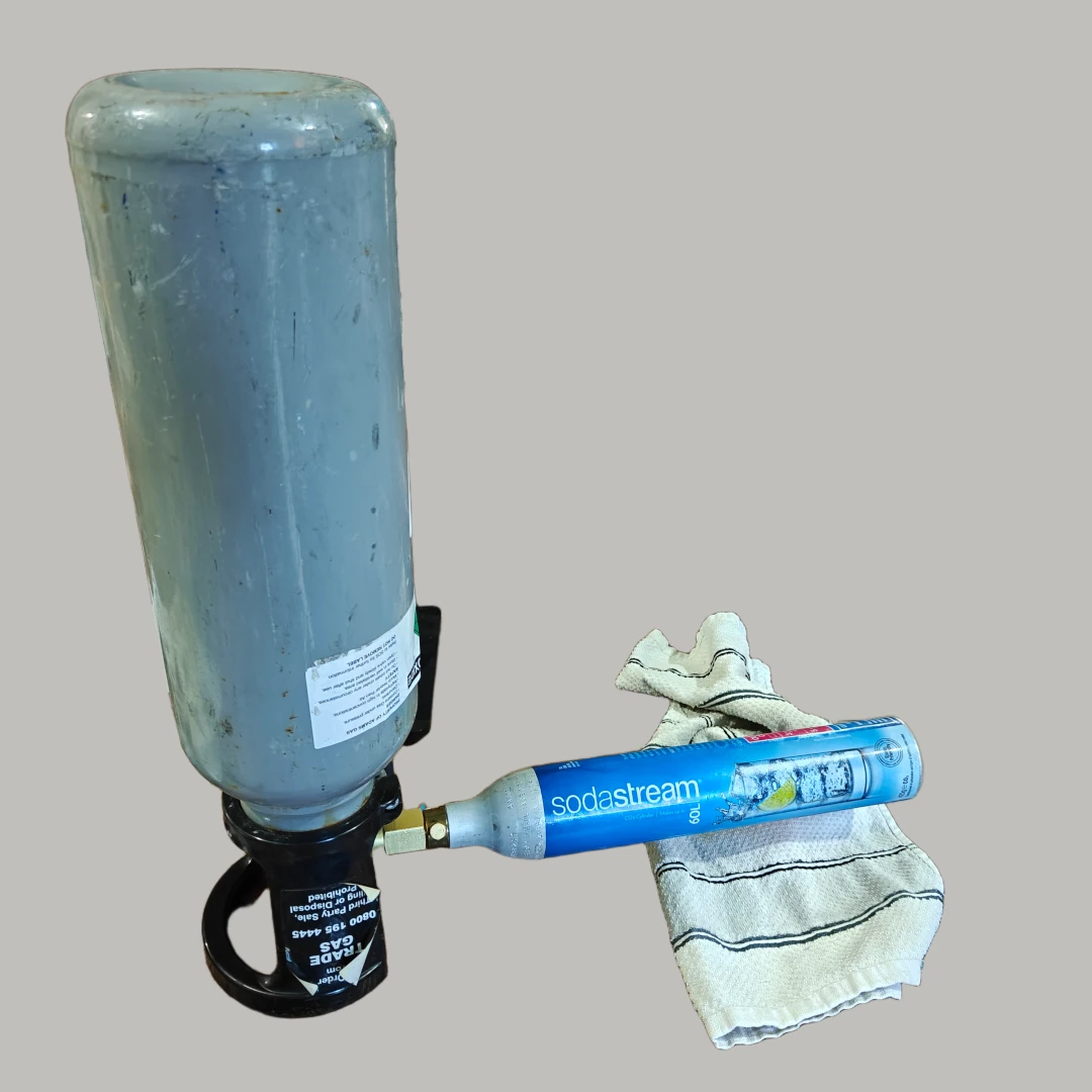 A large gray CO2 bottle sat upside down, with a standard blue smaller Sodastream bottle connected to it via a small straight brass fitting. There’s a small towel under the Sodastream bottle