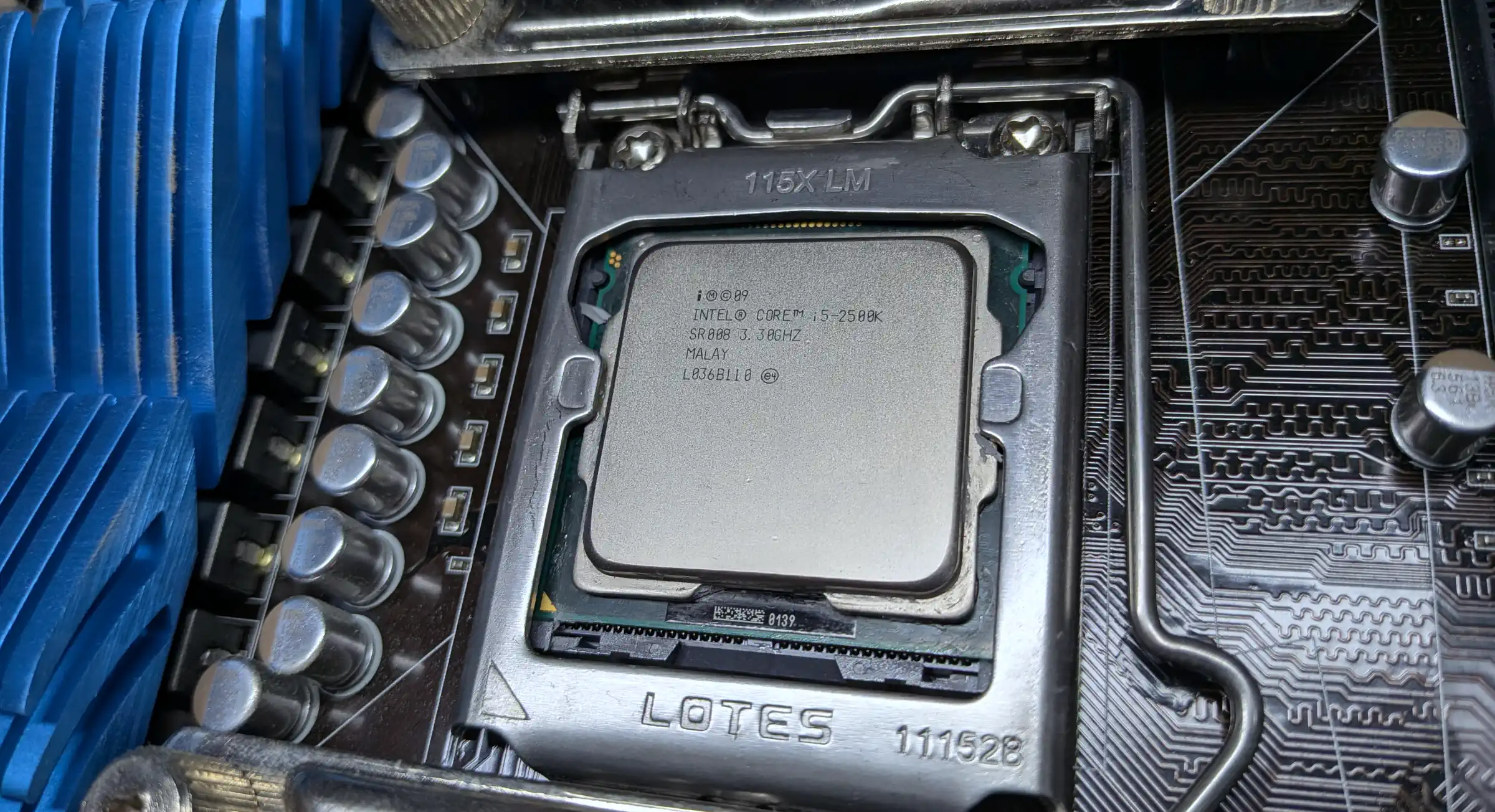 A close up view of a CPU chip, looking like a silver rounded rectangle, in an Intel silver bracket. It&rsquo;s on a brown motherboard with some blue heat sinks visible